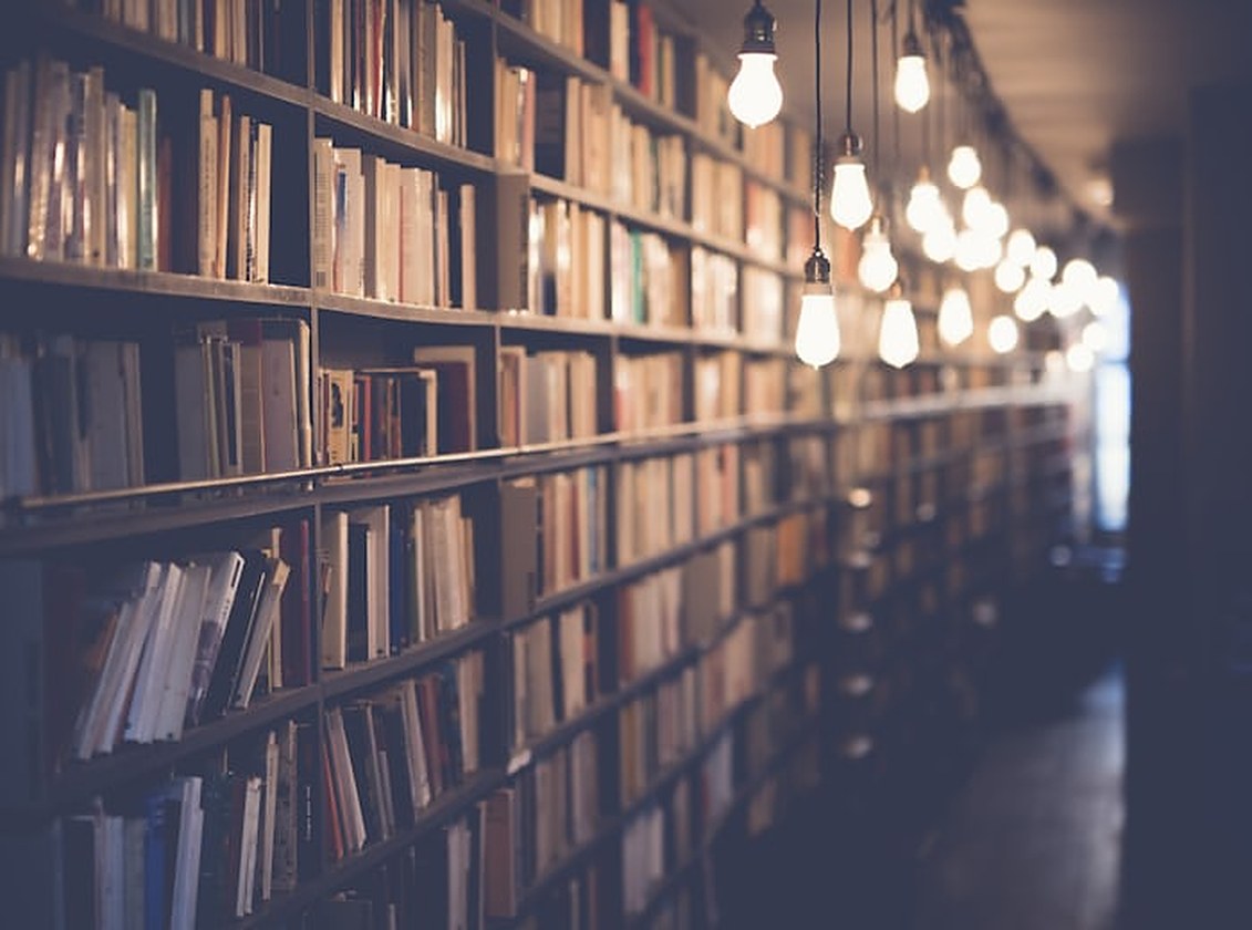 Passadís d’una biblioteca amb prestatgeries plenes de llibres a banda i banda i una filera de bombetes penjants il·luminant el corredor
